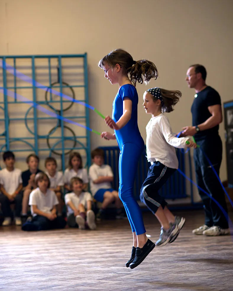 Two girls skip in a PE lesson while their class watches and a skip-hop coach helps