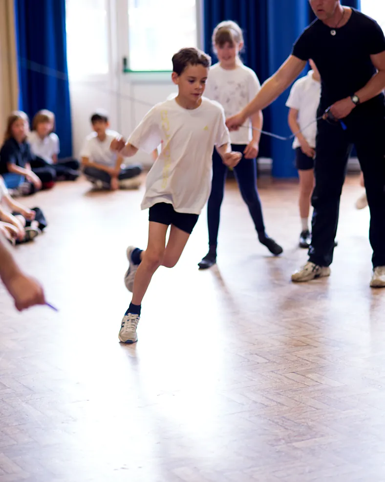 A skip-hop coach guides a pupil running into a long rope in the school hall