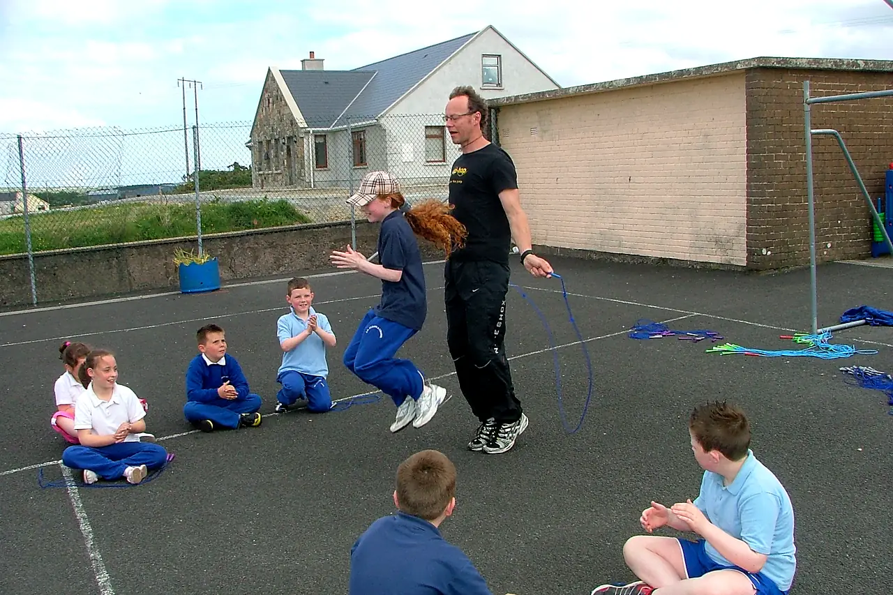 A skip-hop coach demonstrates skipping to a whole class sitting in the school playground