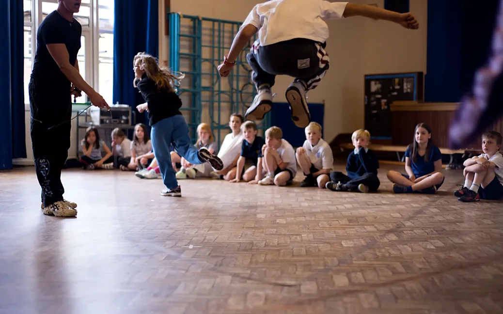 Children skip in a school hall during a skip-hop workshop while classmates watch